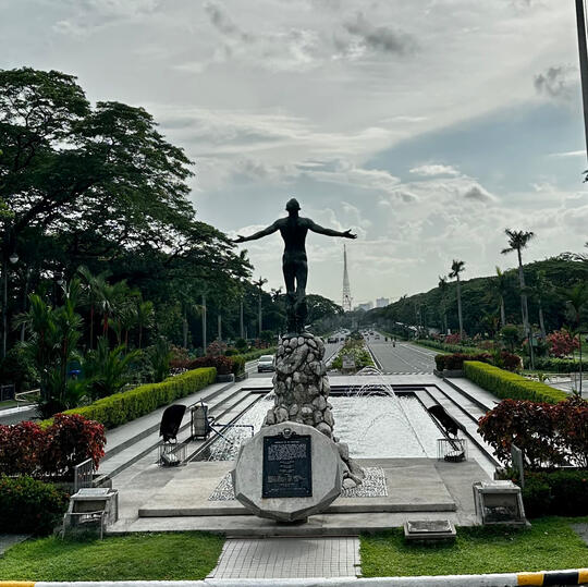 View from the back of the University of the Philippines' Oblation statue, a statue of a naked man facing the sky with arms outreached indicating total submission of self. In front of The Oblation is the plaza with fountain and a road lined with trees